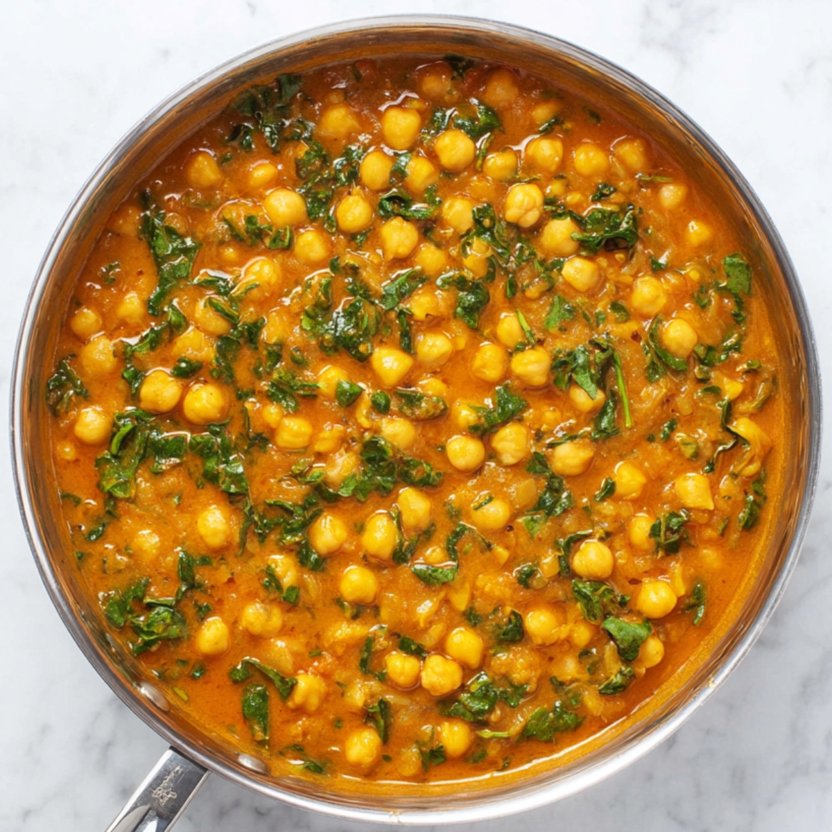 Delicious Chickpea Curry Recipe 13 A stainless steel pan filled with simmering chickpea curry, featuring chickpeas and chopped spinach in a thick, orange sauce. The curry is bubbling on the stove with a clean, white marble background.