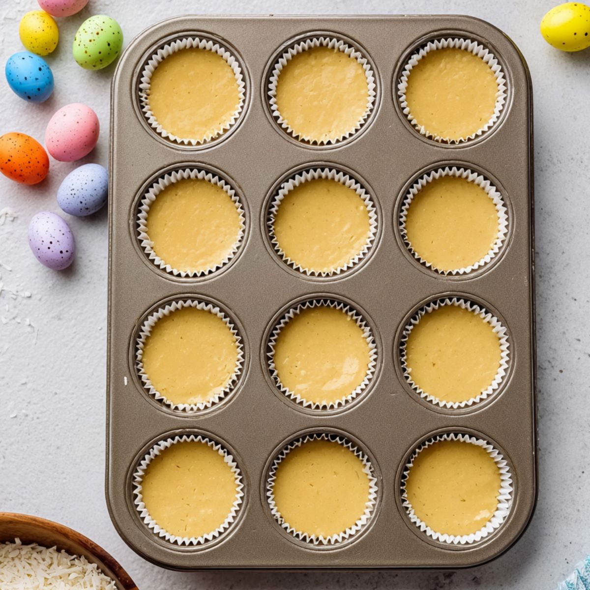 Healthy Coconut Easter Cupcakes Recipe 11 A tray of cupcakes with a light, golden batter before baking, set in paper liners, with a bowl of shredded coconut and colorful Easter candy eggs nearby.