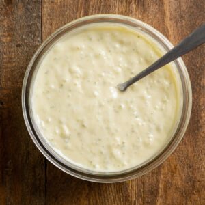 Healthy Coleslaw Recipe Recipe 15 Top-down view of a glass bowl containing creamy coleslaw dressing with a spoon resting inside, placed on a wooden table.