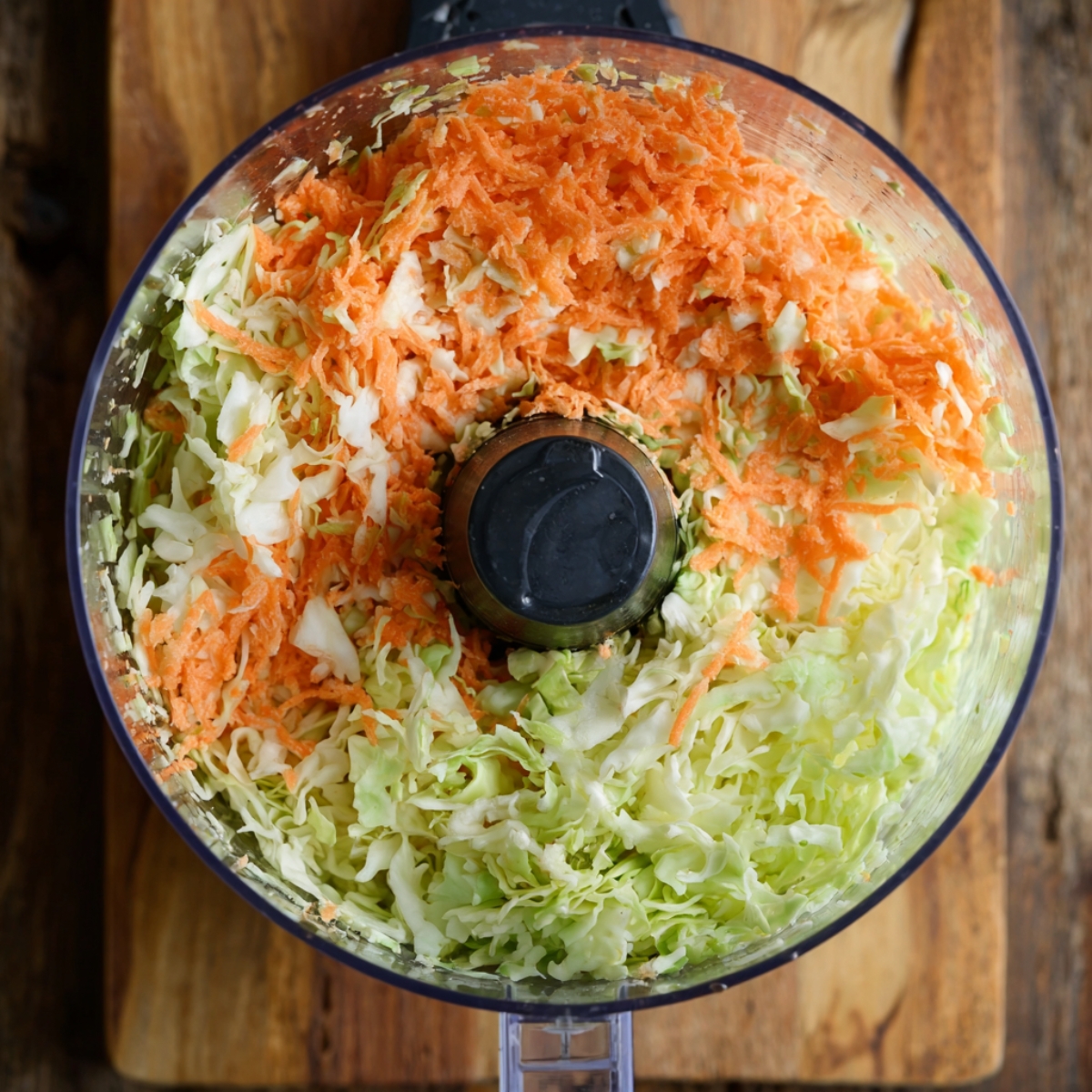 Healthy Coleslaw Recipe Recipe 10 Overhead view of a food processor bowl filled with freshly shredded cabbage and carrots for coleslaw on a wooden surface.