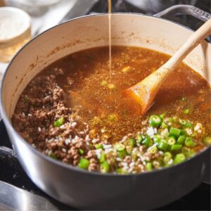 "Rice and ground beef simmering in a pot as broth is being added, wooden spoon stirring, green peppers and spices visible, Moroccan comfort food, warm tones, realistic kitchen scene, high detail, documentary-style food photography."