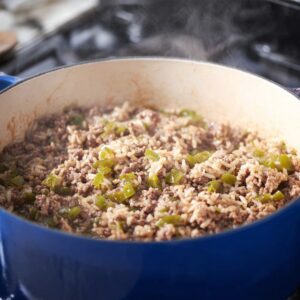 "Ground beef and rice cooking together in a large blue Dutch oven on a stovetop, chopped green peppers visible, steam rising, home-cooked meal, realistic cooking process, natural kitchen lighting, high realism, food photography."