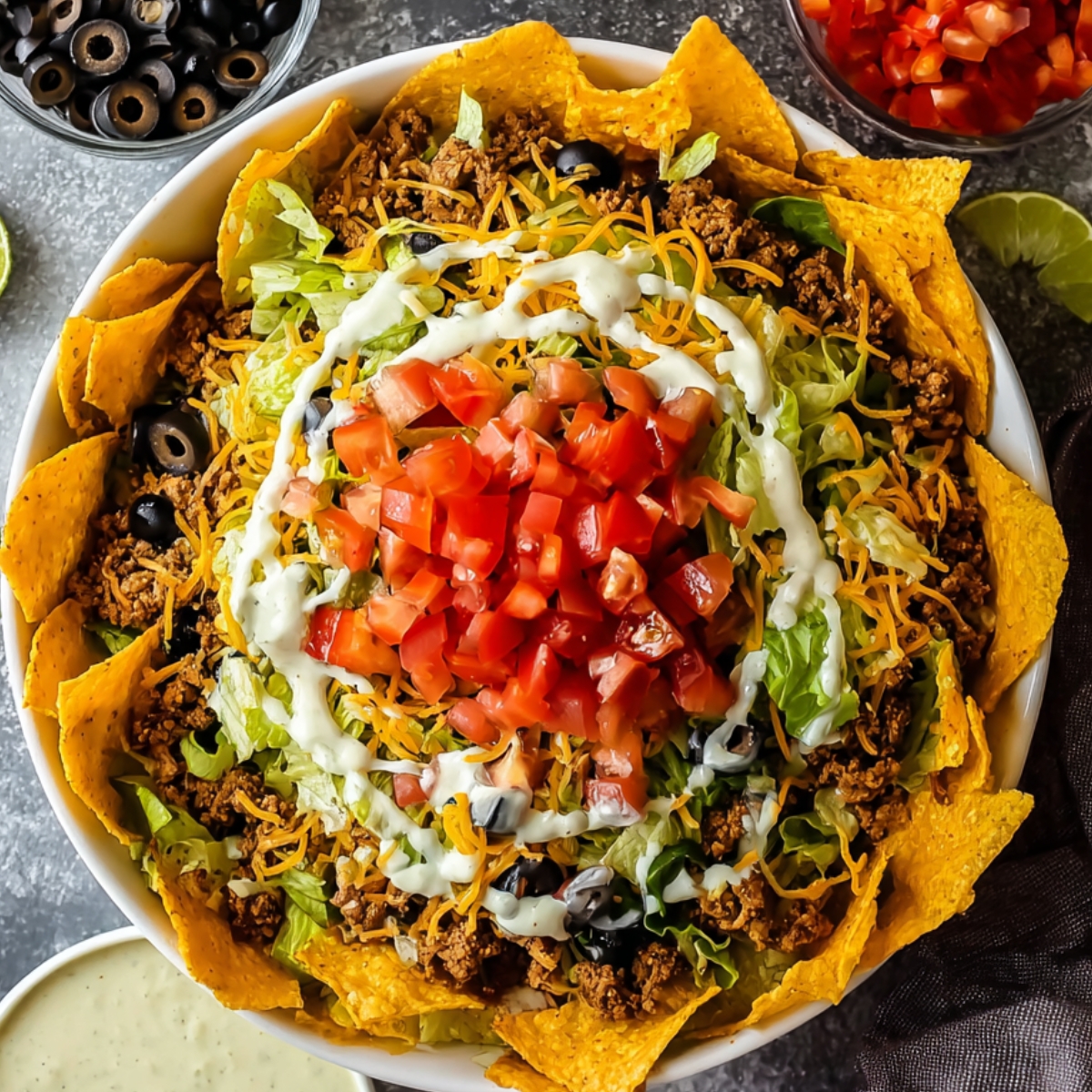 The Best Dorito Taco Salad Recipe 12 A colorful Dorito Taco Salad in a white dish, featuring layers of seasoned ground beef, black beans, chopped tomatoes, black olives, shredded cheese, and a dollop of guacamole. Lime wedges and a few Dorito chips garnish the salad