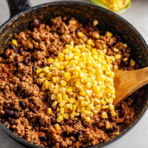 The Best Dorito Taco Salad Recipe 14 Ground beef cooking in a pan, with golden corn kernels being added on top, ready to be mixed into the Dorito Taco Salad for added flavor and texture.