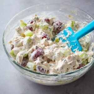 The Best Fruity Chicken Salad Recipe 14 Close-up of creamy chicken and grape salad being mixed in a clear glass bowl with a blue spatula, showing halved red grapes and diced celery in a thick white dressing.