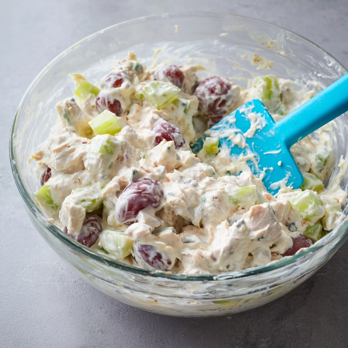 The Best Fruity Chicken Salad Recipe 11 Close-up of creamy chicken and grape salad being mixed in a clear glass bowl with a blue spatula, showing halved red grapes and diced celery in a thick white dressing.