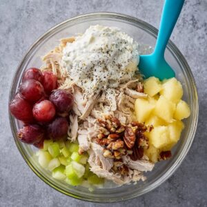 The Best Fruity Chicken Salad Recipe 13 Overhead view of chicken salad ingredients in a glass bowl, including shredded chicken, mayonnaise, sour cream, red grapes, chopped celery, crushed pineapple, pecans, and black pepper before mixing.