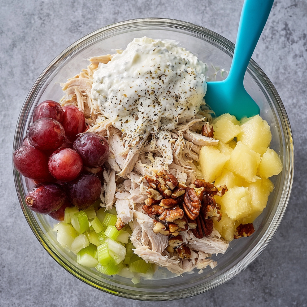 The Best Fruity Chicken Salad Recipe 10 Overhead view of chicken salad ingredients in a glass bowl, including shredded chicken, mayonnaise, sour cream, red grapes, chopped celery, crushed pineapple, pecans, and black pepper before mixing.