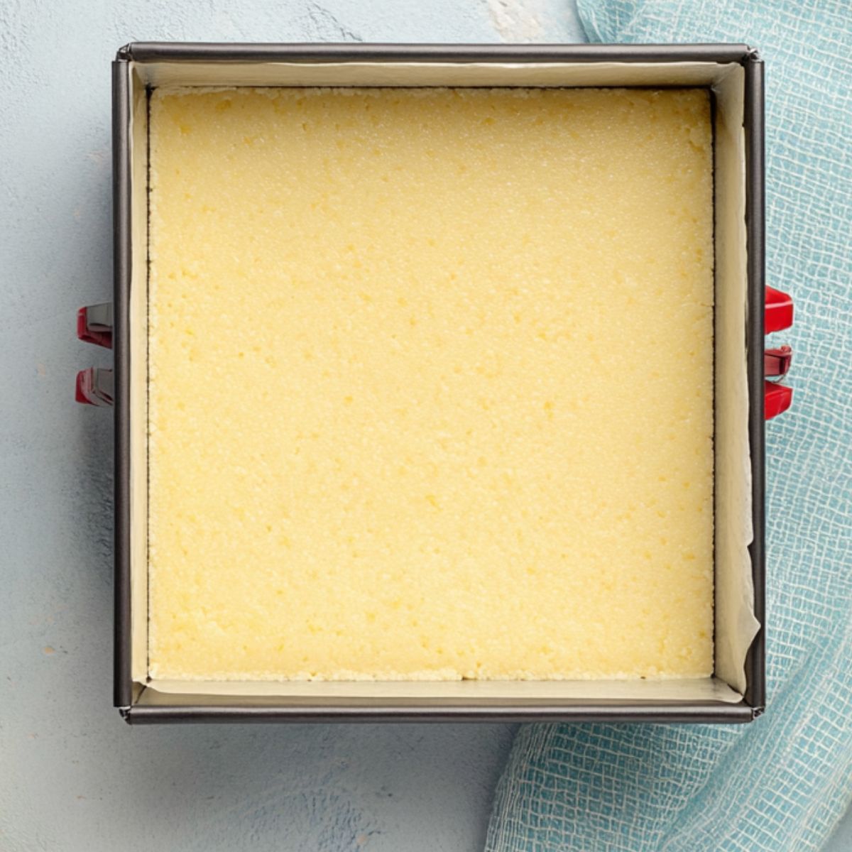 Delicious Lemon Bars Recipe 11 A square baking pan with raw lemon bar dough inside, ready to be baked, surrounded by fresh lemons and a light blue checkered cloth."