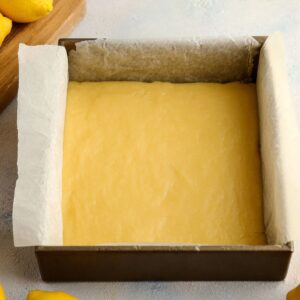Delicious Lemon Bars Recipe 16 A square pan filled with a smooth, golden lemon bar mixture ready to be cut into squares, with parchment paper hanging from the edges, surrounded by fresh lemons.