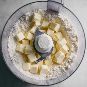 Easy Lemon Blueberry Tart Recipe 15 A food processor filled with flour and small cubes of butter, ready to be mixed into a dough. The butter is placed on top of the flour, and the food processor blade is centered in the mixture, about to begin processing the ingredients