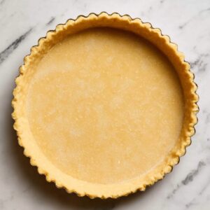 Easy Lemon Blueberry Tart Recipe 16 A raw, unbaked tart crust resting in a tart pan. The crust has a golden color and a scalloped edge, ready for baking. The marble surface below the tart adds a nice touch to the presentation.