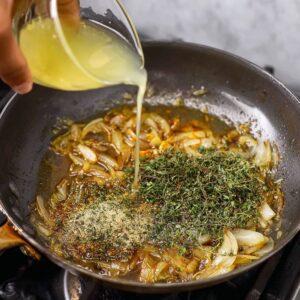 The pan is enhanced with fresh herbs and a splash of liquid, creating a fragrant base for the sauce. As the liquid melds with the caramelized onions and herbs, it begins to form the foundation of a flavorful glaze that will coat the chicken