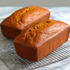 The Best Pumpkin Bread Recipe 18 Two freshly baked pumpkin bread loafs cooling on a wire rack, with golden-brown crusts and soft interiors, ready to be sliced.