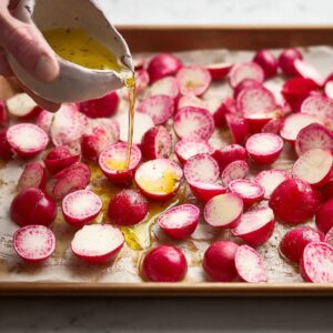 Easy Roasted Radishes Recipe 15 "A hand pouring olive oil over halved radishes, which are spread out on a parchment-lined baking sheet, preparing them for roasting. The vibrant red color of the radishes contrasts with the golden oil."
