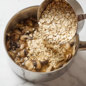 An overhead cooking process shot of rolled oats being poured from a metal measuring cup into a saucepan with mushrooms and broth, mid-action pour, white marble countertop, bright natural light, sharp focus, minimalistic kitchen style.