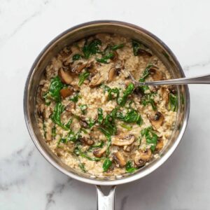 A top-down view of a stainless steel saucepan filled with creamy savory oatmeal mixed with sautéed mushrooms and fresh spinach, spoon inside the pot, clean white marble background, soft natural lighting, realistic food photography.