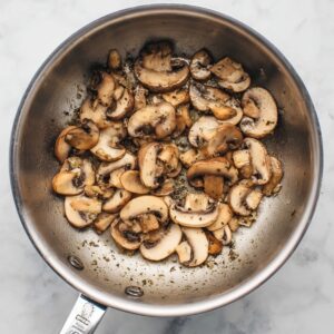 A top-down view of sliced mushrooms being sautéed in a stainless steel saucepan, lightly browned, simple cooking scene, white marble background, natural lighting, realistic texture, clean and minimal food photography.