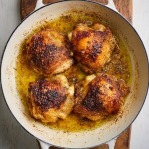 The Best Smothered Chicken Recipe 16 "Golden-brown chicken thighs with crispy skin cooking in a white enamel pan, surrounded by sizzling juices and seasoning. The chicken looks flavorful and perfectly seared on both sides.