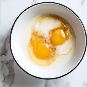 Best Sourdough Chocolate Chip Scones 16 Top-down view of a white ceramic bowl with a black rim on a white marble countertop, containing two raw eggs, visible egg yolks and egg whites mixed with vanilla extract, soft natural lighting, minimal food photography, high resolution, clean background, realistic details