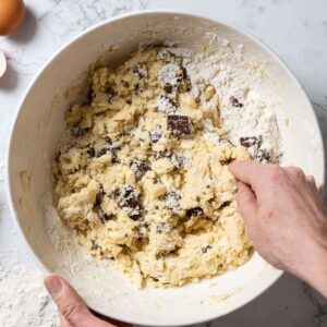 Best Sourdough Chocolate Chip Scones 17 Overhead shot of hands mixing chocolate chip scone dough in a white bowl, flour and wet ingredients partially combined, visible chocolate chunks, marble countertop background, natural daylight, step-by-step baking process, realistic food photography, high resolution