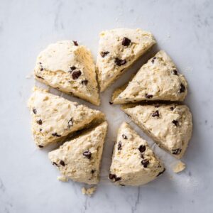 Best Sourdough Chocolate Chip Scones 18 Top view of raw chocolate chip scone dough cut into triangular pieces, thick and soft dough with visible chocolate chunks, placed on parchment paper, minimal white marble background, natural light, food preparation process, high detail, realistic kitchen photography
