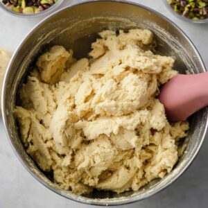 A mixing bowl of plain spumoni cookie dough, unflavored and smooth, with a spatula resting inside, ready to be divided and flavored into pistachio, cherry, and chocolate dough components for spumoni cookies.