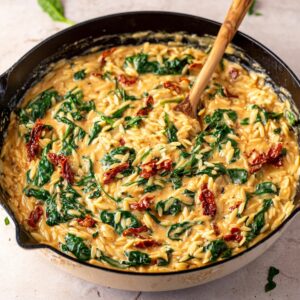 A creamy skillet with orzo pasta, spinach, and sun-dried tomatoes mixed together. The dish has a smooth, rich sauce, and the spinach is wilted, creating a vibrant, comforting one-pan meal.