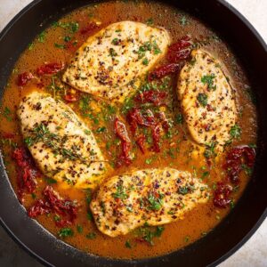 Chicken breasts cooking in a skillet, surrounded by a flavorful broth with sun-dried tomatoes and herbs. The chicken is seasoned and browning in the pan, ready to be simmered with the other ingredients.