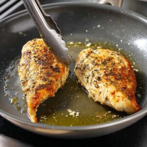 A skillet with golden-brown chicken breasts resting on creamy orzo pasta with sun-dried tomatoes and spinach. The dish is garnished with fresh herbs, creating a beautiful, hearty meal.