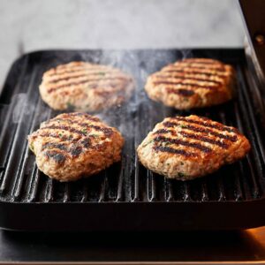 Delicious Grilled Chicken Burgers Recipe 18 The chicken patties are being grilled on a stovetop or grill, showing grill marks, which indicates they’re cooking to perfection for that crispy exterior and juicy interior.