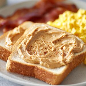 Delicious Cinnamon Butter Recipe 16 Close-up of two slices of toast spread thickly with cinnamon honey butter on a white plate, with scrambled eggs and crispy bacon blurred in the background.