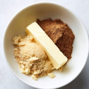 Delicious Cinnamon Butter Recipe 14 A white bowl on a light surface containing a stick of butter, brown sugar, and ground cinnamon piled together but not yet mixed.