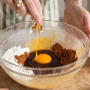 A person cracking an egg into a mixing bowl containing other ingredients, possibly for a gingerbread cookie dough.