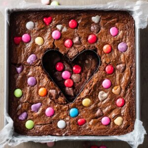 Healthy Heart Shaped Brownies Recipe 18 A pan of brownies topped with colorful candy-coated chocolate pieces, with a heart-shaped cookie cutter in the center creating a fun shape.