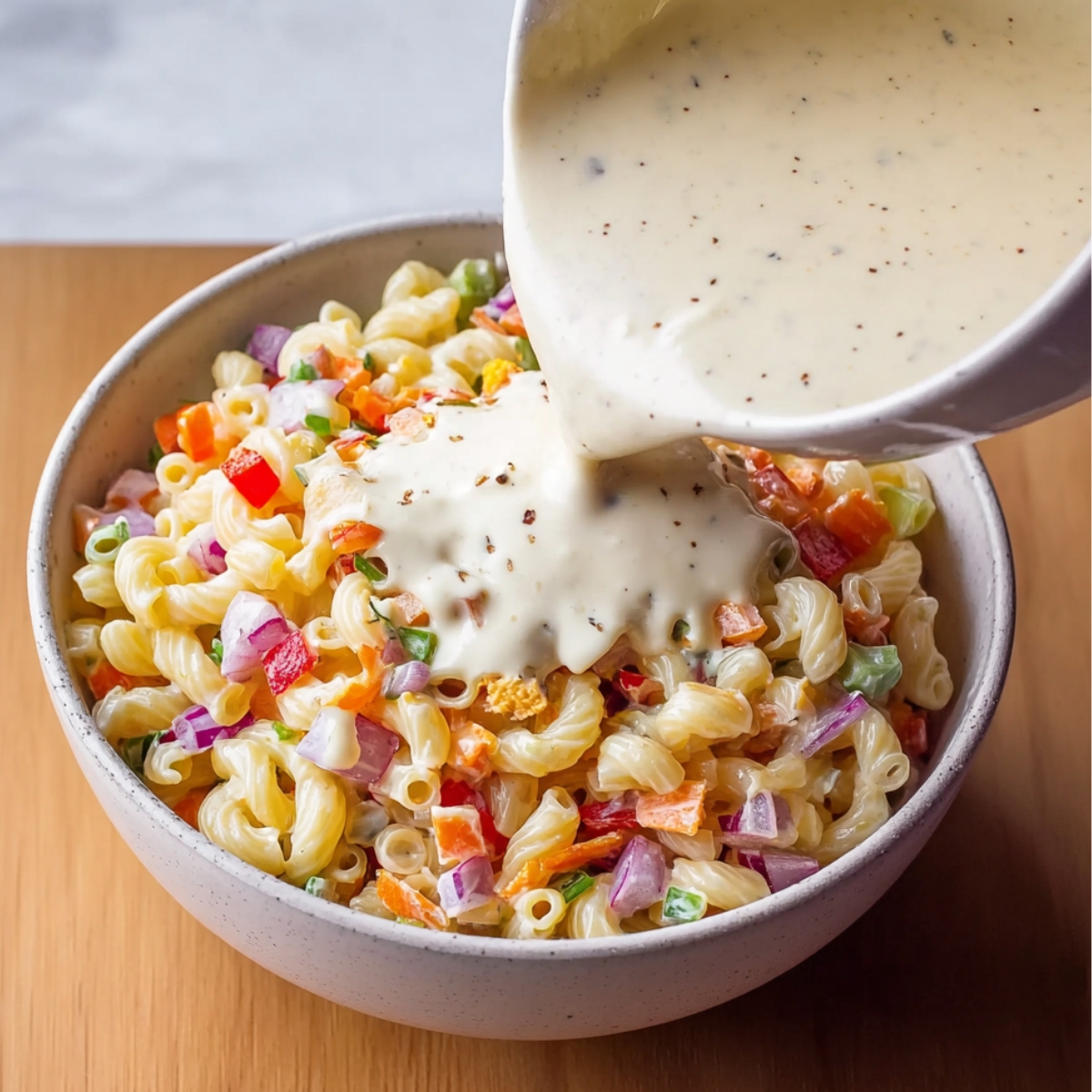 Best Macaroni Salad Recipe 13 Pouring creamy dressing over the macaroni salad filled with chopped vegetables. The dressing is being added to bring all the flavors together in a smooth, rich texture