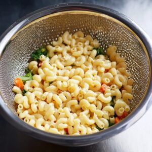 Strained cooked macaroni in a colander, ready to be mixed with the vegetables and dressing for the perfect macaroni salad. The pasta is properly drained to avoid a watery salad."
