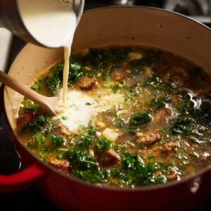 Easy Zuppa Toscana Soup Recipe 15 A close-up of cream being poured into a simmering pot of Zuppa Toscana, filled with kale, sausage, and potatoes in a creamy broth.