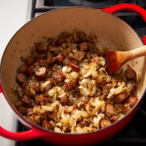 Easy Zuppa Toscana Soup Recipe 14 Ground sausage and onions being sautéed in a red pot on the stovetop, with the ingredients beginning to brown and release their aroma.