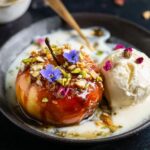 Baked Peaches A plated dessert featuring a glossy caramelized apple topped with nuts and edible flowers, served alongside a scoop of vanilla ice cream drizzled with a creamy sauce on a dark plate.