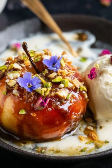 The Best Baked Peaches Recipe 1 Baked Peaches A plated dessert featuring a glossy caramelized apple topped with nuts and edible flowers, served alongside a scoop of vanilla ice cream drizzled with a creamy sauce on a dark plate.