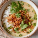 Ginger Miso Brothy Soup A bowl of miso-ginger brothy soup served with white rice, topped with crispy shredded ingredients, fresh cilantro leaves, and sliced green onions. The soup is creamy and light beige in color, and the bowl has a rustic white interior with a brown rim, placed on a beige cloth.
