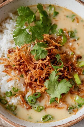 Best Ginger Miso Brothy Soup Recipe 1 Ginger Miso Brothy Soup A bowl of miso-ginger brothy soup served with white rice, topped with crispy shredded ingredients, fresh cilantro leaves, and sliced green onions. The soup is creamy and light beige in color, and the bowl has a rustic white interior with a brown rim, placed on a beige cloth.