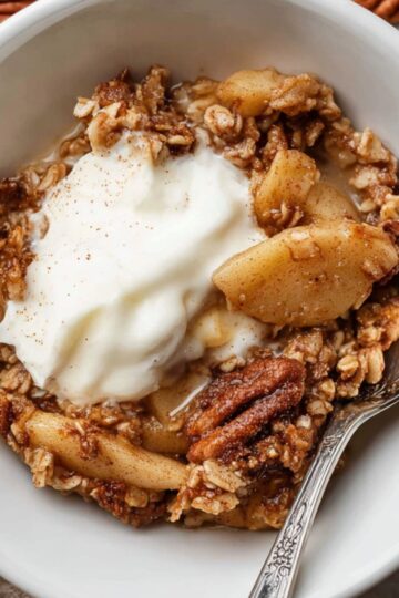 Best Baked Apple Oatmeal Recipe 1 baked apple oatmeal A serving of baked apple oatmeal in a white bowl, topped with a scoop of yogurt and a spoon, with pecans scattered around.