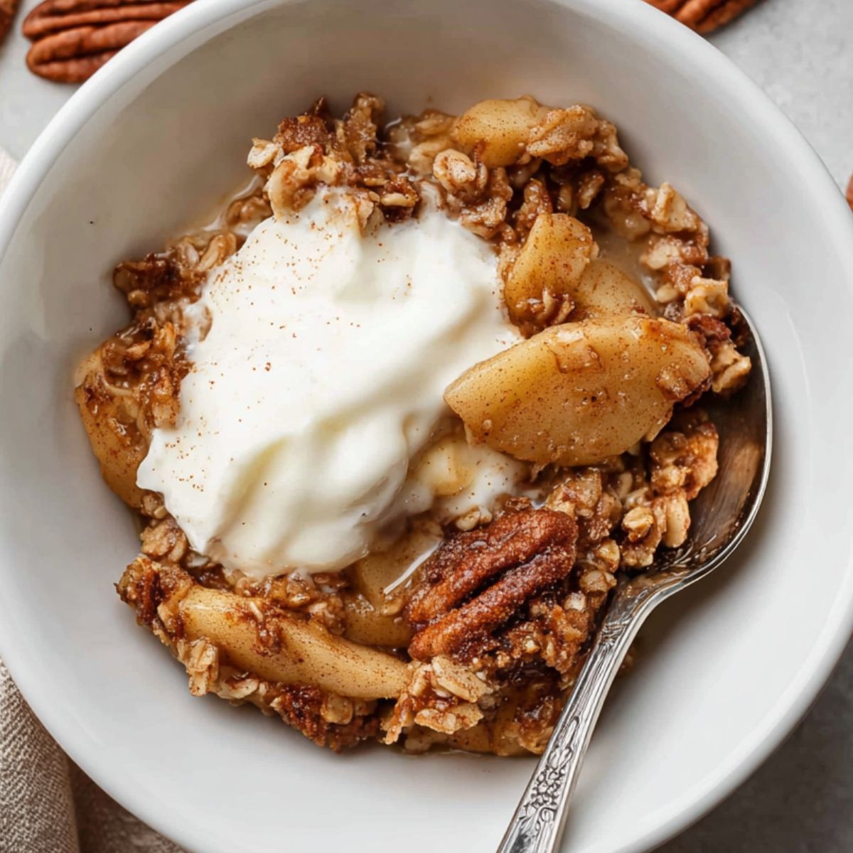 baked apple oatmeal A serving of baked apple oatmeal in a white bowl, topped with a scoop of yogurt and a spoon, with pecans scattered around.