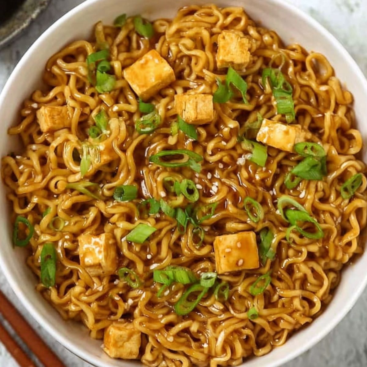 Bowl of saucy ramen noodles garnished with green onions and tofu cubes, ready to eat, with chopsticks on the side.