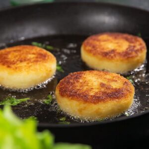 Fried potato cheese pancakes cooking in a skillet, with golden crispy edges.