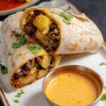Black Bean Burritos A freshly made wrap sliced in half, revealing a vibrant filling of roasted potatoes, black beans, and creamy sauce, served with a side of spicy sauce and garnished with fresh cilantro for an added touch.
