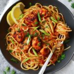 Blackened Shrimp Pasta A plate of spaghetti with tomato sauce and seasoned chicken pieces, garnished with parsley and green onions, served with a lemon slice on the side and a fork in the dish.