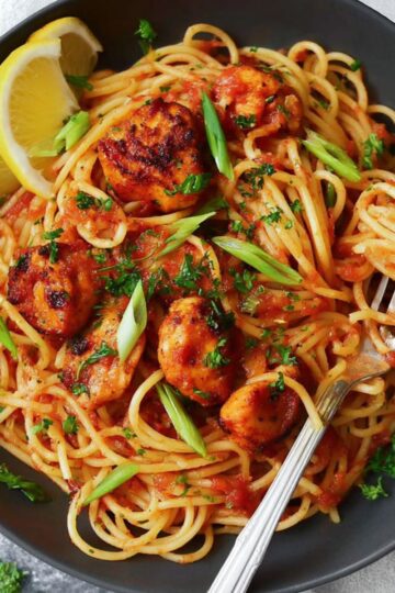 Healthy Blackened Shrimp Pasta Recipe 1 Blackened Shrimp Pasta A plate of spaghetti with tomato sauce and seasoned chicken pieces, garnished with parsley and green onions, served with a lemon slice on the side and a fork in the dish.